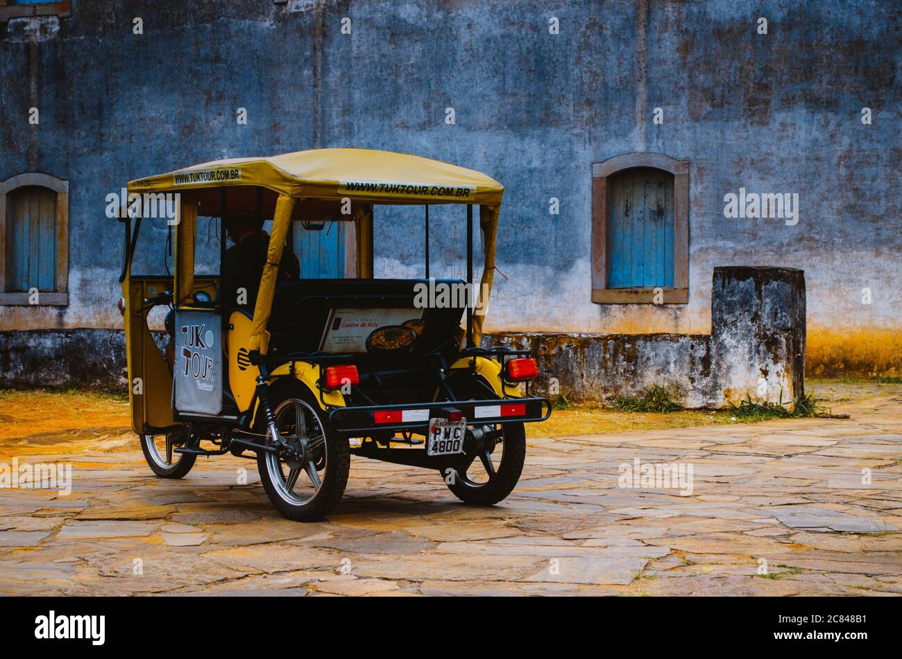 Ein gelbes Tuk-Tuk-Fahrzeug der Firma Tuk Tour vor einer alten Steinmauer aus einer touristischen Kirche in Ouro Preto, Minas Gerais. Stockfoto