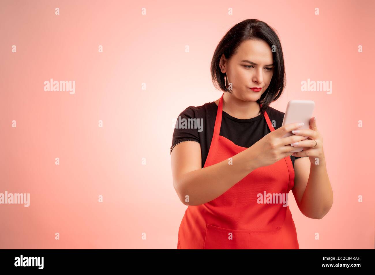 Frau im Supermarkt mit roter Schürze und schwarzem T-Shirt beschäftigt, hält Smartphone, SMS, nervös isoliert auf rotem Hintergrund Stockfoto