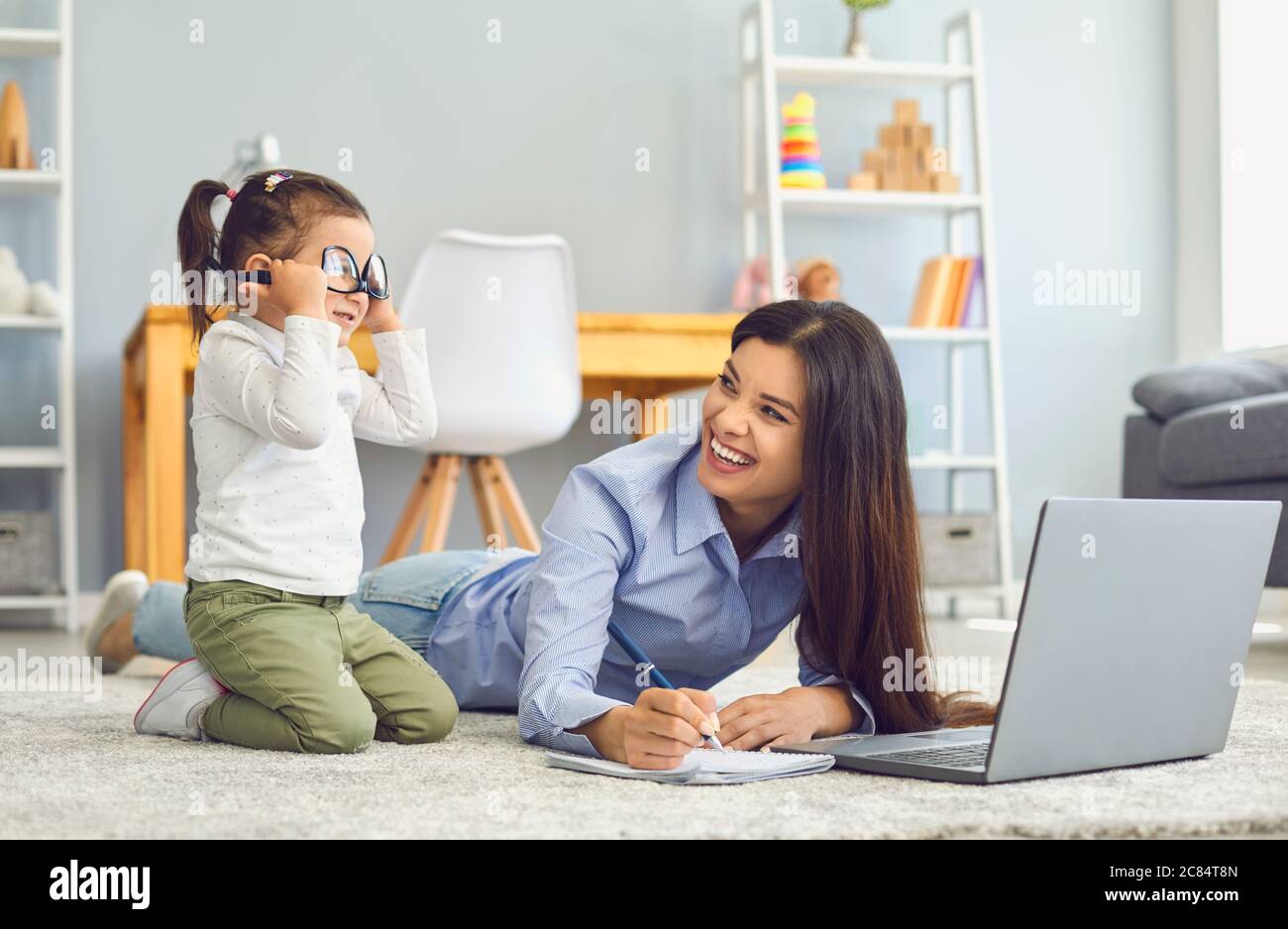 Liebenswert Mädchen spielen mit Brille, während sie Mutter tun online freiberuflich zu Hause. Arbeiten von zu Hause und Elternschaft Stockfoto
