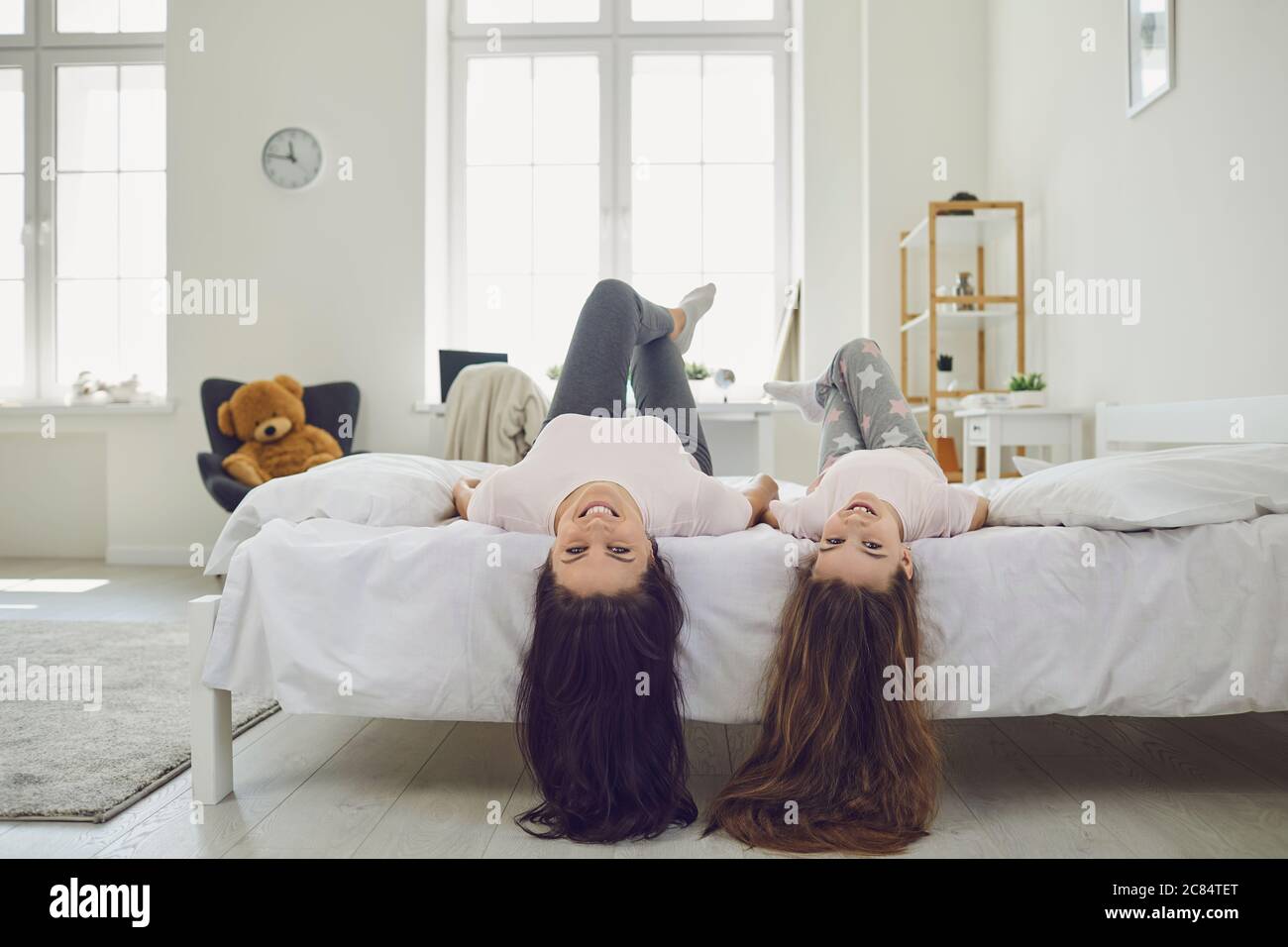 Fröhliche Mutter und Tochter mit schönen Haaren, die Kopf nach unten auf dem Bett zu Hause liegen. Eltern und Kinder haben Spaß im Schlafzimmer Stockfoto
