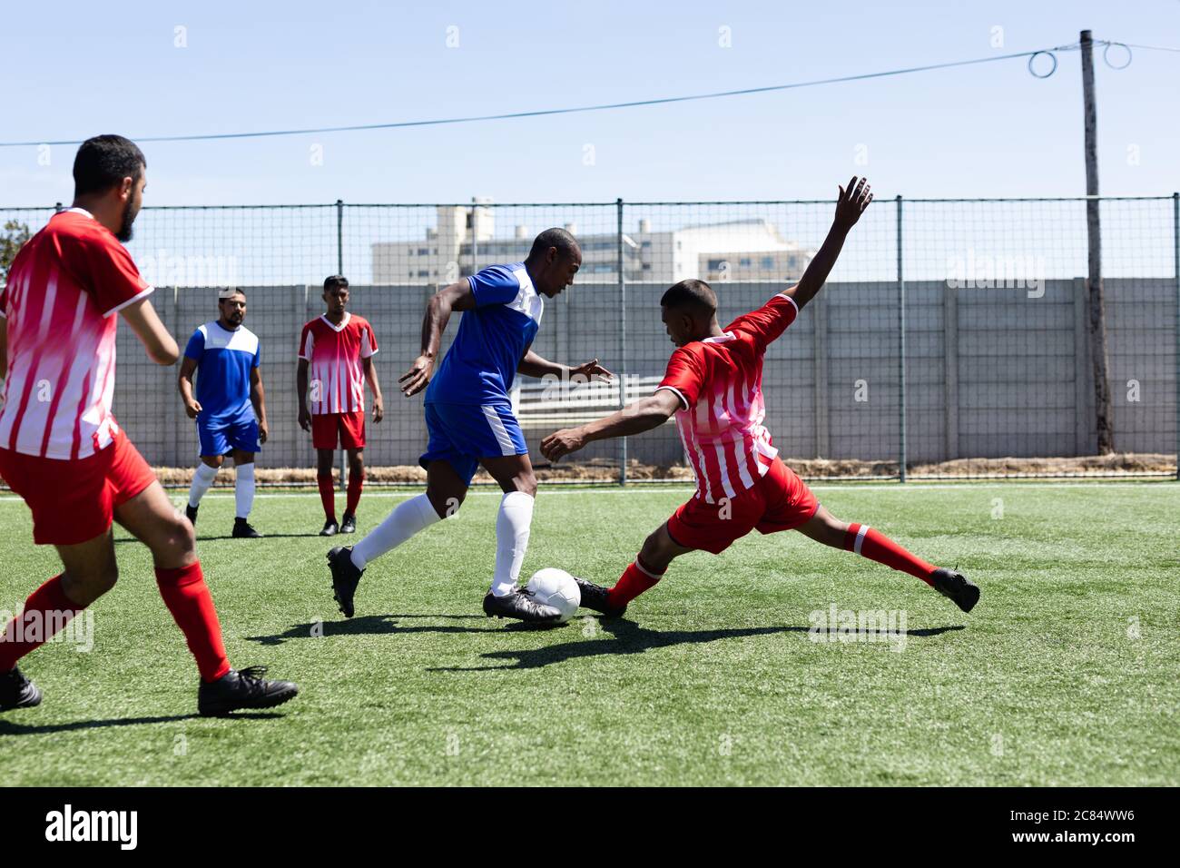 Zwei multiethnische Mannschaften von männlichen fünf A-Side-Fußballspieler tragen einen Mannschaftsstreifen spielen ein Spiel auf einem Sportplatz in der Sonne, Tackling und tritt bal Stockfoto