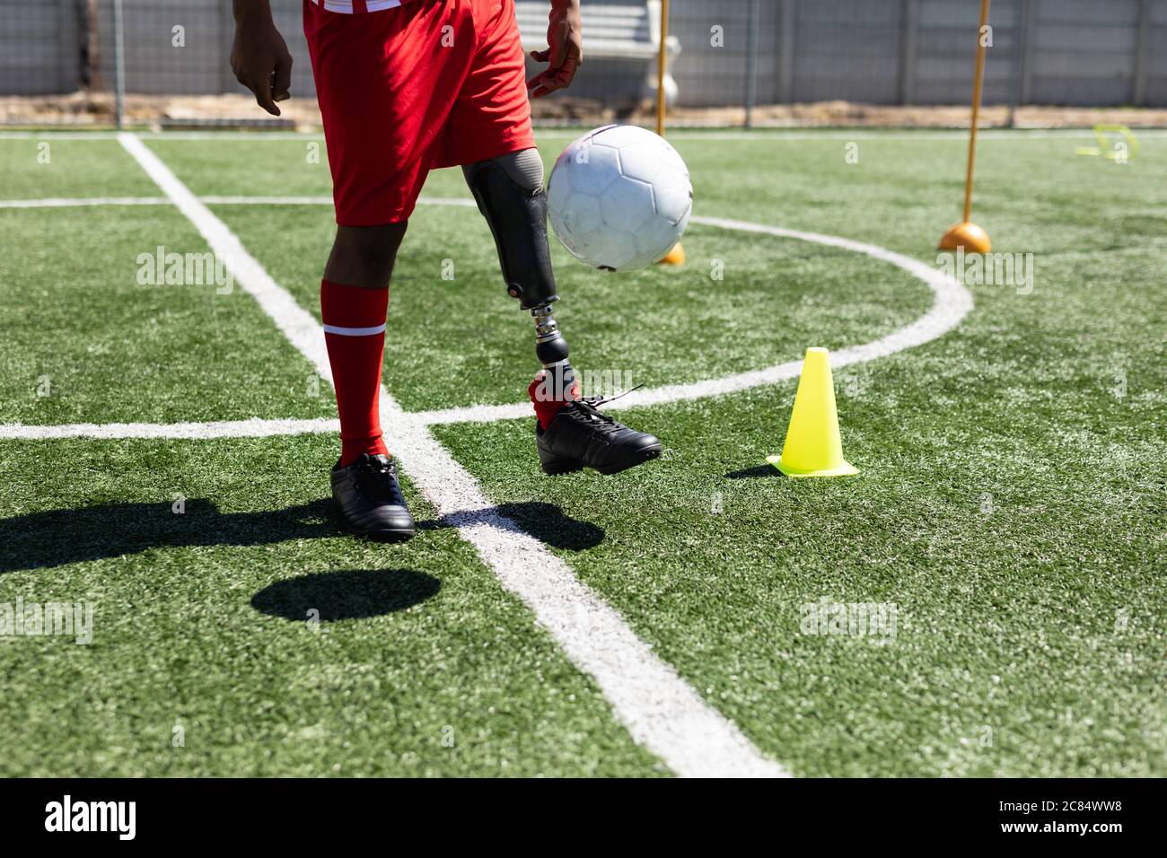 Gemischter Fußballspieler mit Beinprothese, der einen Mannschaftsstreifen trägt, der auf einem Sportplatz in der Sonne trainiert und den Ball aufwärmt. Stockfoto