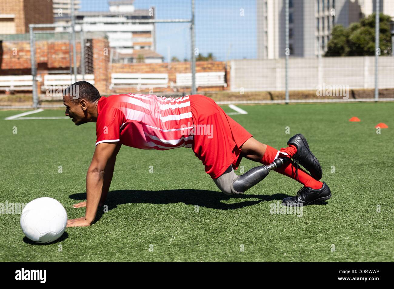 Gemischter Fußballspieler mit Beinprothese, der ein Mannschaftsstreifen trägt, der auf einem Sportplatz in der Sonne trainiert, und sich dabei aufwärmt, Liegestütze mit Ball ne zu machen Stockfoto