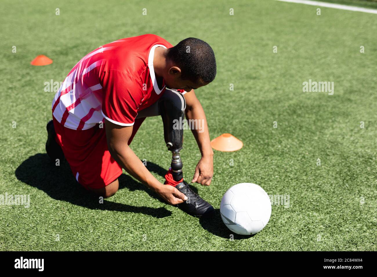 Gemischter Fußballspieler mit Beinprothese, der einen Mannschaftsstreifen trägt, der auf einem Sportplatz in der Sonne trainiert und neben ihm einen Schnürsenkel bindet. Stockfoto