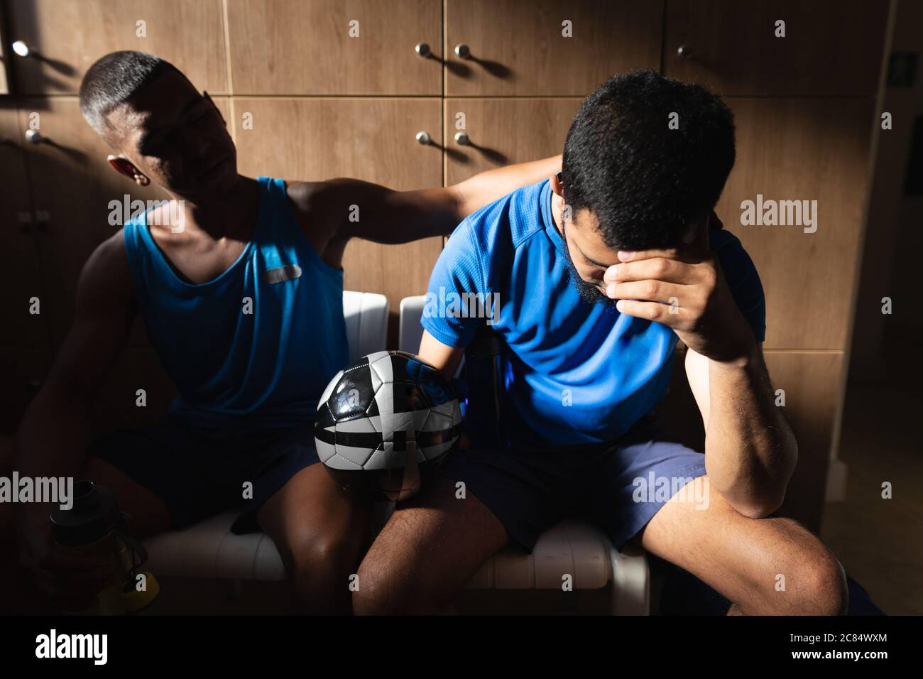 Zwei Männer mit gemischtem Rennen, die in Sportkleidung in der Umkleidekabinen sitzen, während einer Spielpause, den Ball in Ruhe halten und enttäuscht sein. Stockfoto