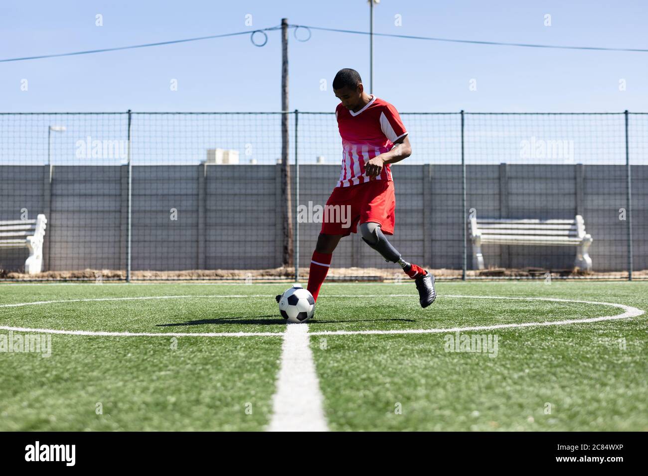 Gemischter Fußballspieler mit Beinprothese, der einen Mannschaftsstreifen trägt, der auf einem Sportplatz in der Sonne trainiert und den Ball aufwärmt. Stockfoto
