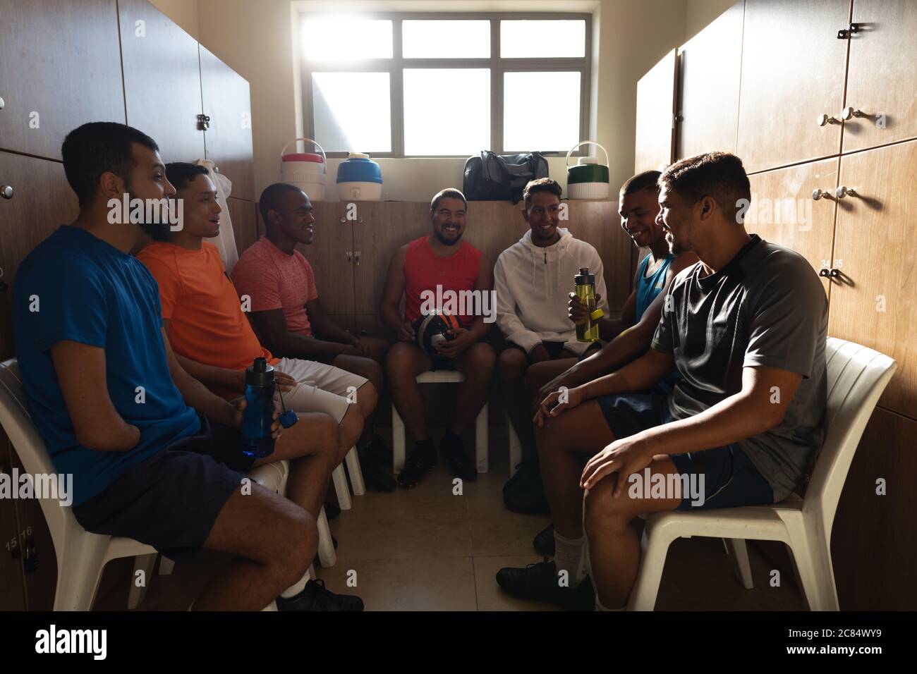 Multiethnische Gruppe von Fußballspielern in Sportkleidung sitzen in Umkleidekabinen während einer Pause im Spiel, halten Ball und Wasserflasche lächelnd t Stockfoto