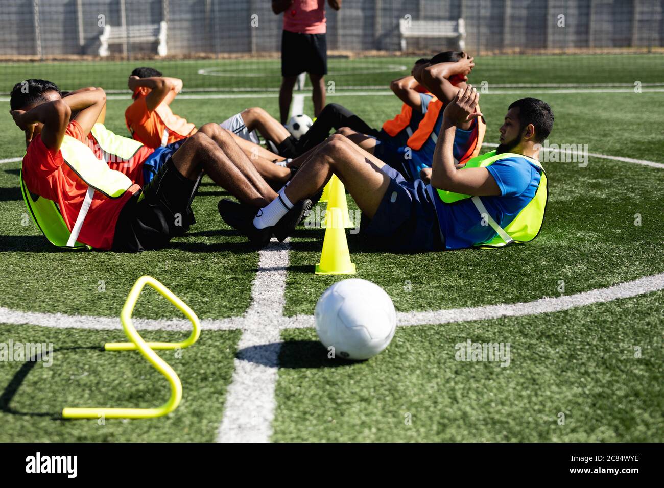 Multi ethnische Gruppe von männlichen fünf ein Seite Fußballspieler tragen Sportkleidung und Westen Training auf einem Sportplatz in der Sonne, Aufwärmen dabei Sit U Stockfoto