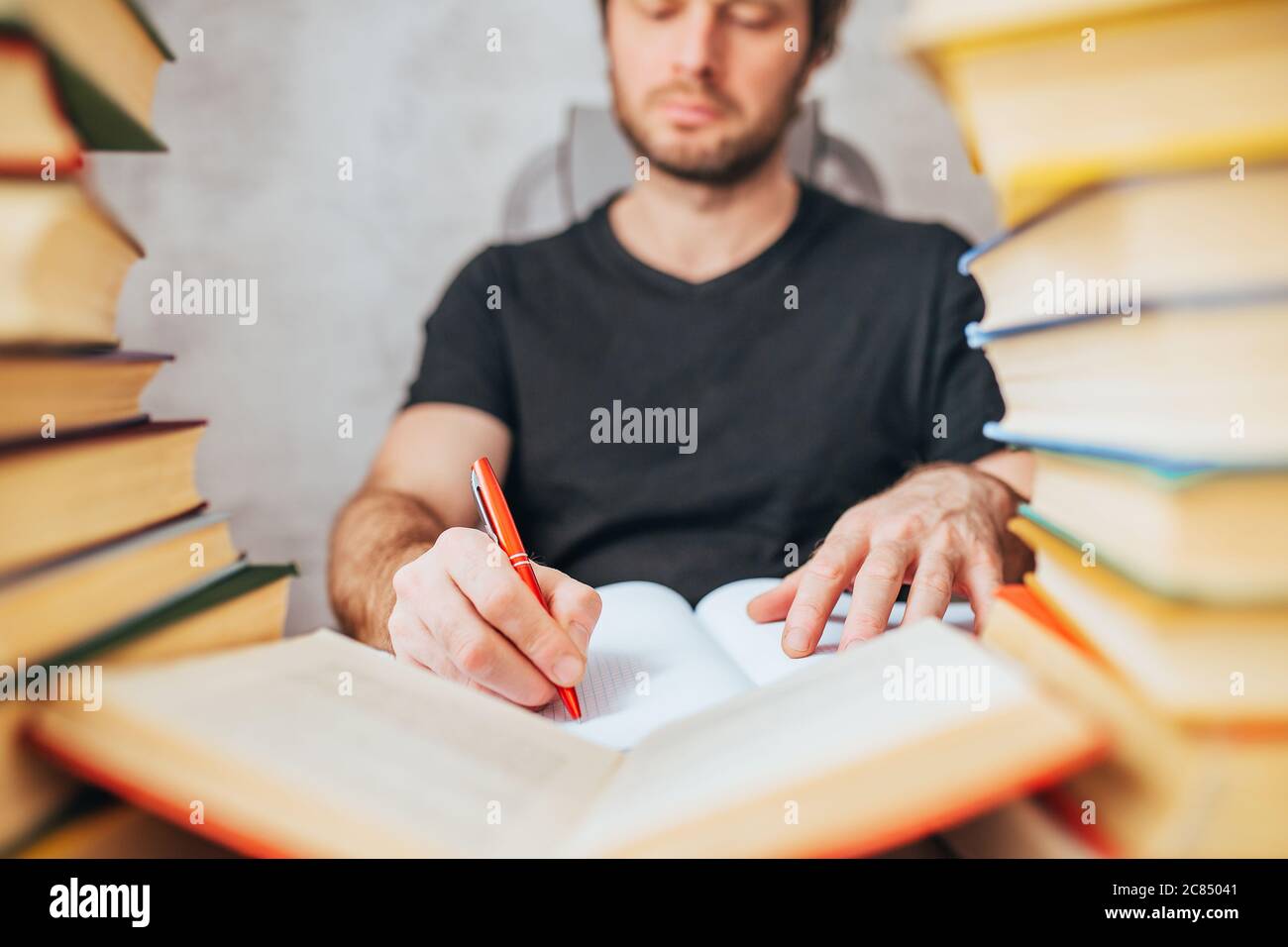 Ein Mann mit Kugelschreiber schreibt in einem Notizbuch in einer Bibliothek, umgeben von Büchern - wissenschaftliche Arbeit Stockfoto