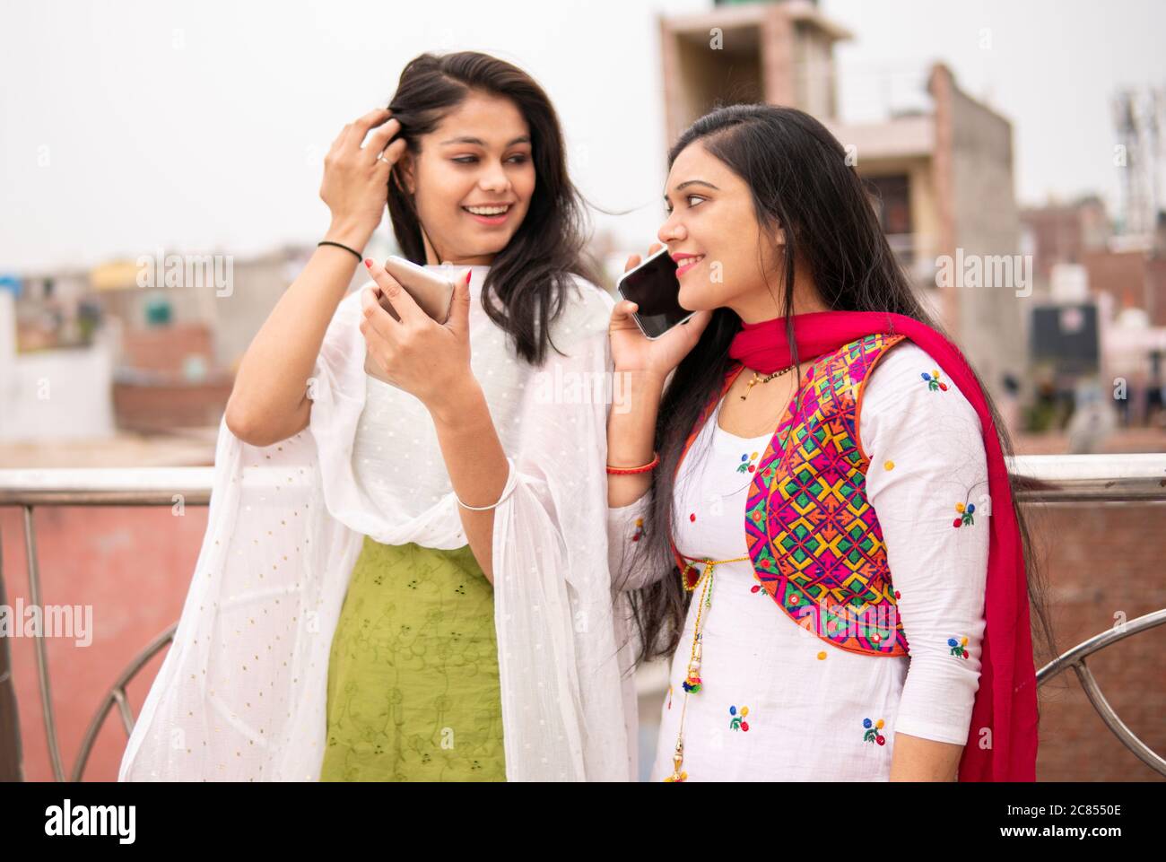 Schöne indische junge Frauen stehen zusammen in der Nähe Geländer. Eine Frau ist mit dem Smartphone beschäftigt und eine andere telefoniert. Stockfoto