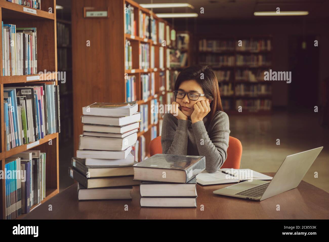 Gelangweilte junge Studentin, die am Tisch in einer offenen Bibliothek auf dem Campus sitzt. Bildungskonzept Stockfoto
