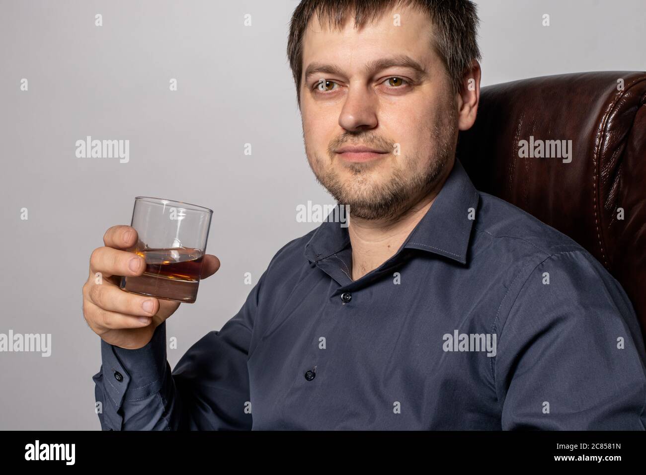 Ein hübscher junger Mann von 30-35 Jahren im Hemd sitzt auf einem braunen Ledersessel und hält ein Glas Whiskey in der Hand. Stockfoto