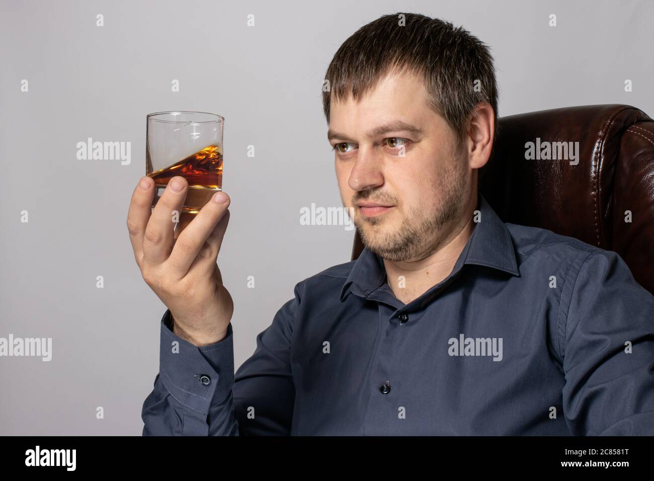 Ein hübscher junger Mann von 30-35 Jahren im Hemd sitzt auf einem braunen Ledersessel und hält ein Glas Whiskey in der Hand. Stockfoto