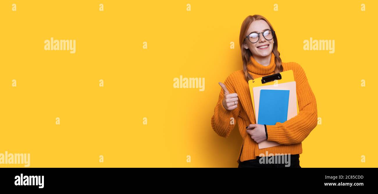 Schöne rothaarige Student mit Sommersprossen gestikulierende die wie Zeichen und hält Ordner an einer gelben Wand mit freiem Platz Stockfoto