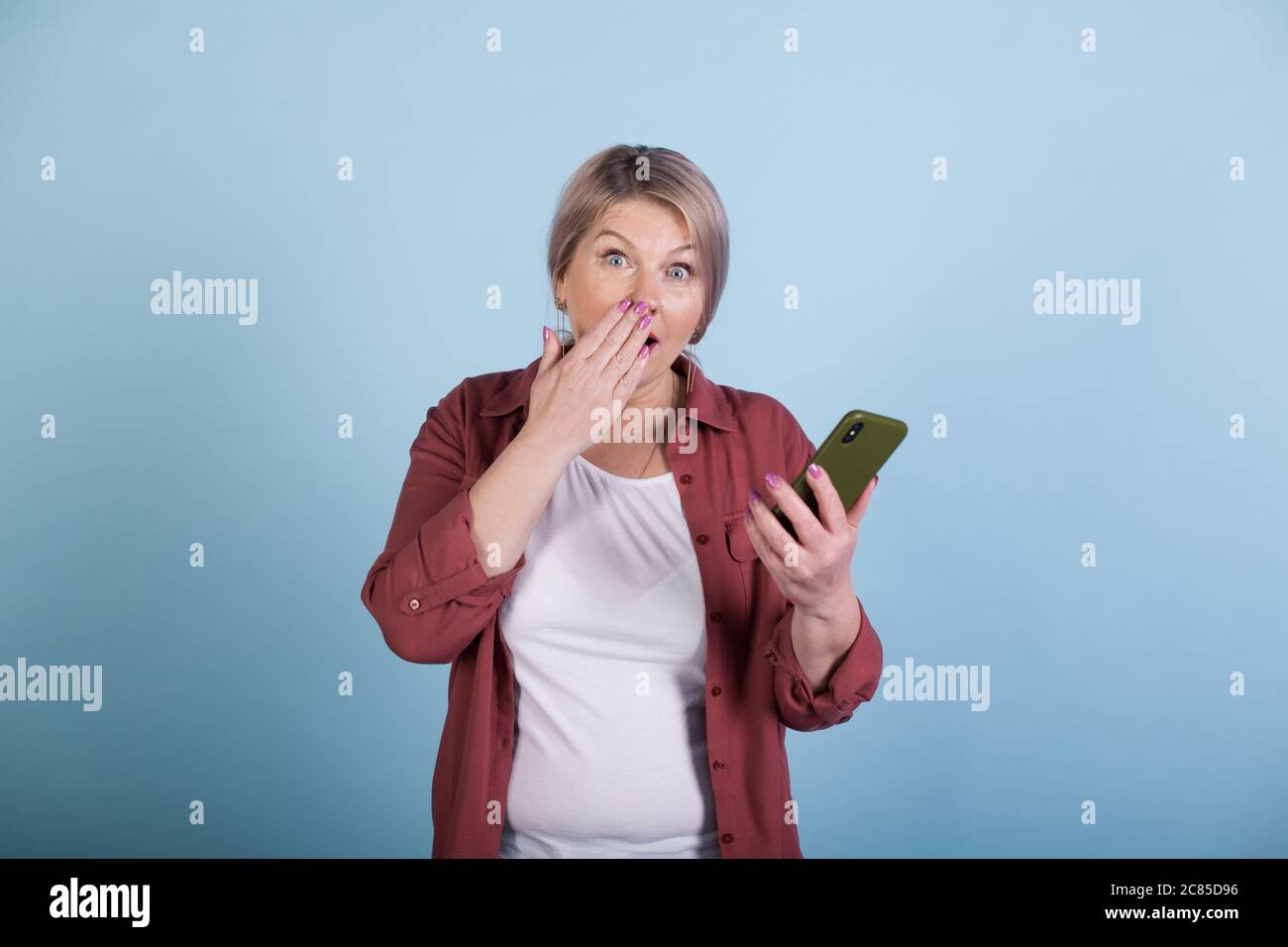 Schockierte kaukasische ältere Frau mit blonden Haaren, die ein Telefon hält und die Kamera an einer blauen Studiowand anschaut Stockfoto