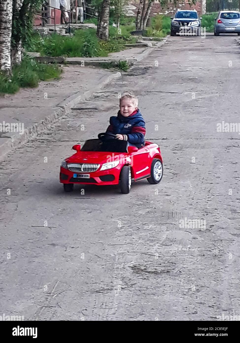 Kleiner weißer Junge von drei Jahren in einem roten Kinder-Elektroauto fährt durch Stadtstraßen - vertikales Foto Stockfoto