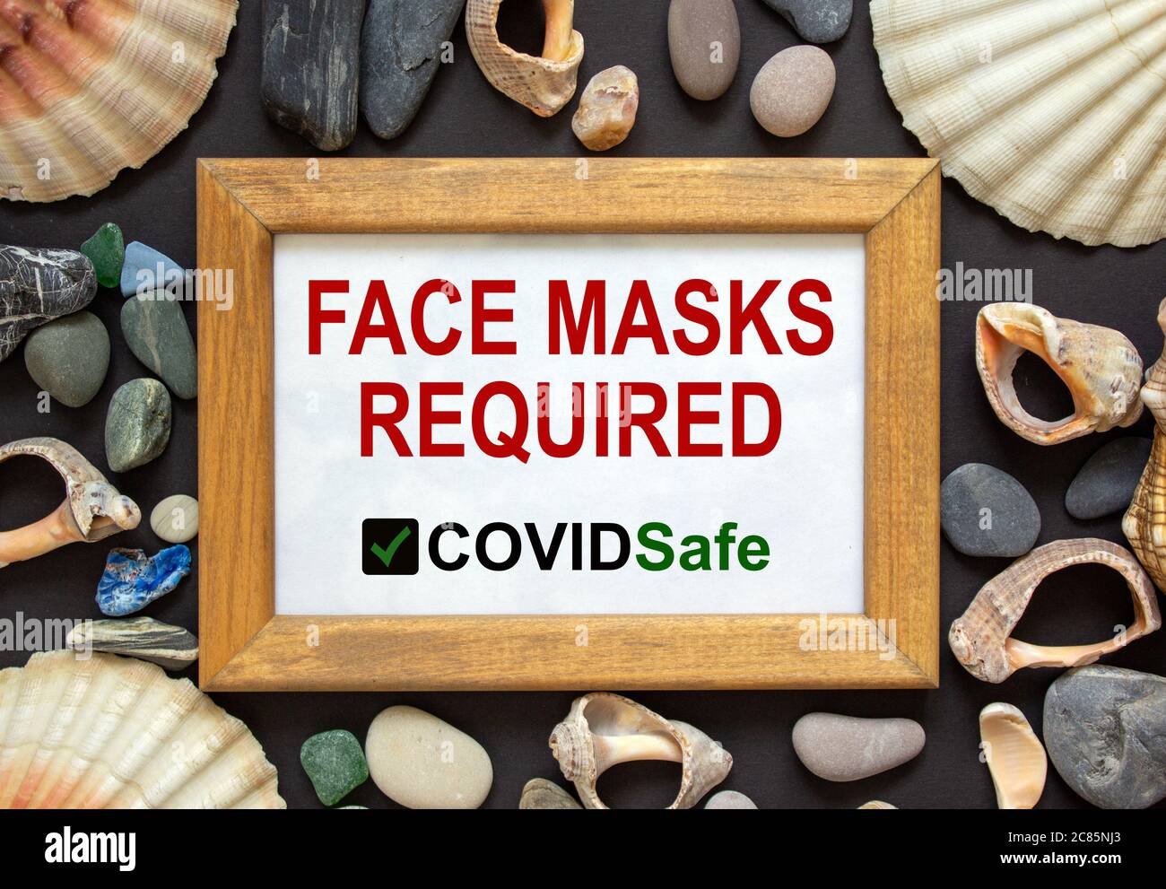 Holzbildrahmen mit Aufschrift "Gesichtsmasken erforderlich covid sicher" auf schönem schwarzen Hintergrund. Seesteine und Muscheln. Konzept. Stockfoto