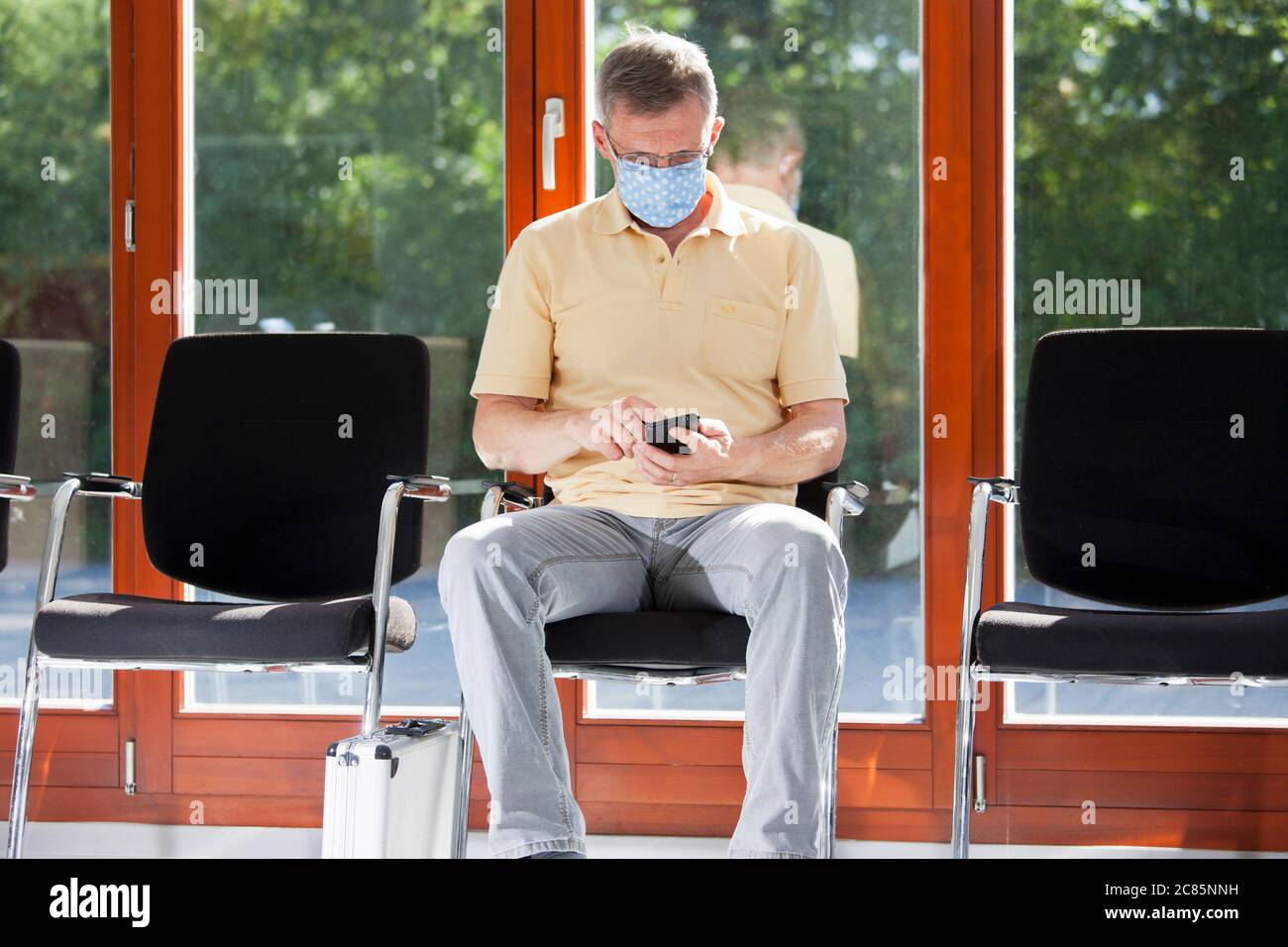 Reifer Mann mit Gesichtsmaske sitzt in einem hellen Wartezimmer eines Krankenhauses oder eines Büros Blick auf Smartphone Stockfoto