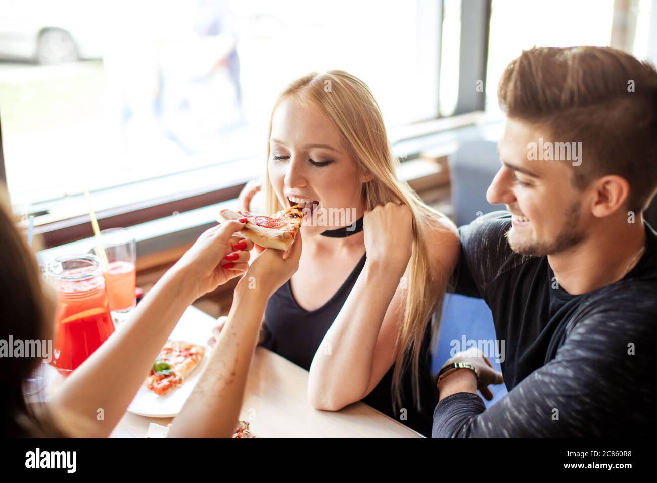 Fröhliche glückliche männliche und weibliche Studenten behandeln einander mit Bio-Pizza mit verschiedenen Belägen und nicht alchoholic Cocktails, Spaß und en Stockfoto