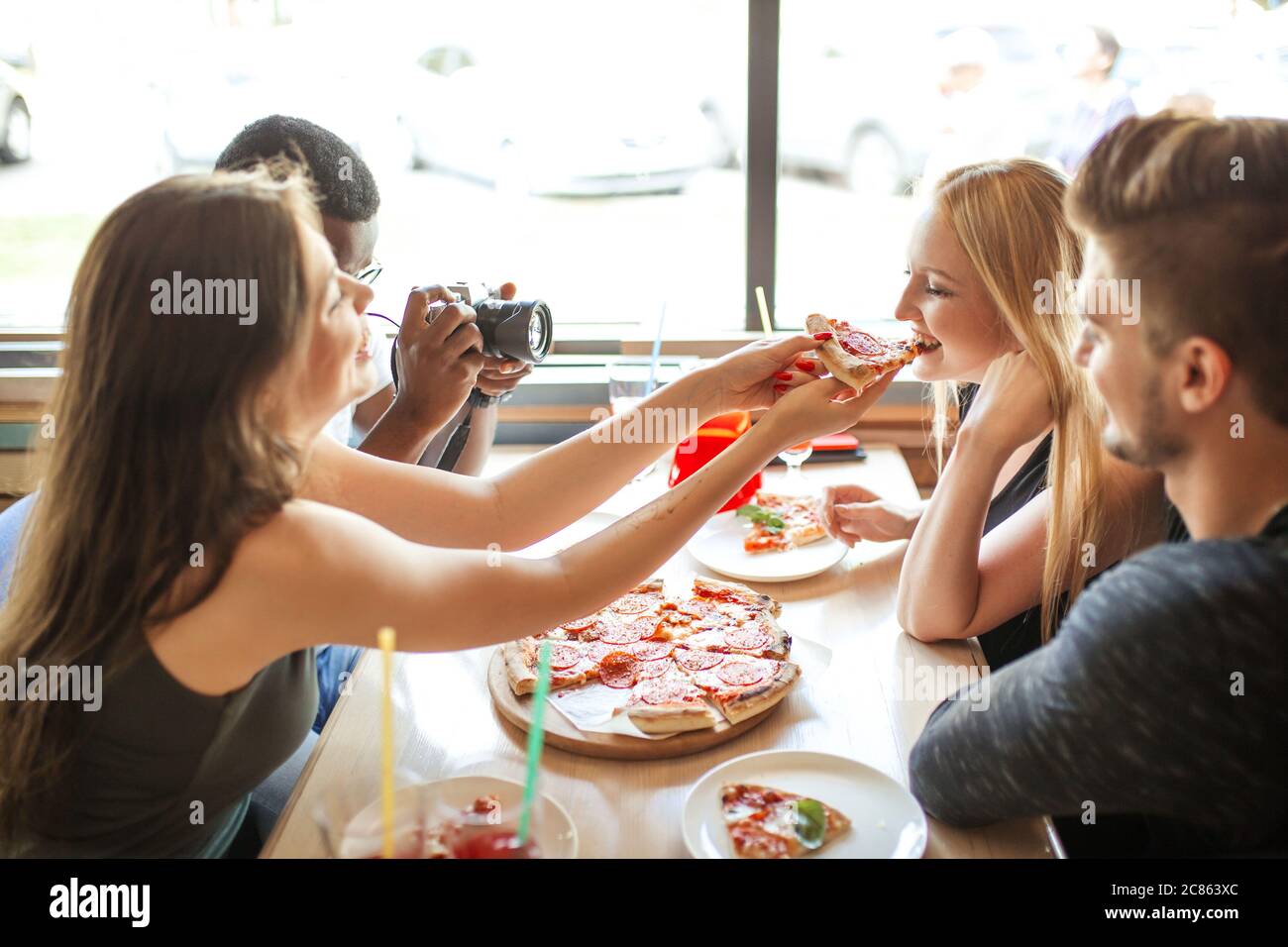 Fröhliche glückliche männliche und weibliche Studenten behandeln einander mit Bio-Pizza mit verschiedenen Belägen und nicht alchoholic Cocktails, Spaß und en Stockfoto