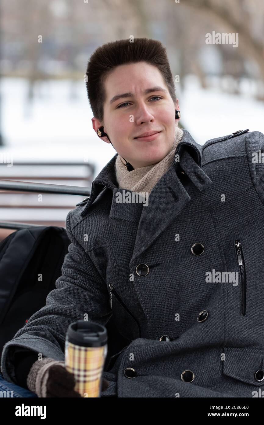 Ein glücklicher Student sitzt auf einer Bank in einem Pavillon im Park, hört Musik über Kopfhörer und trinkt Kaffee aus einer wiederverwendbaren Tasse Stockfoto