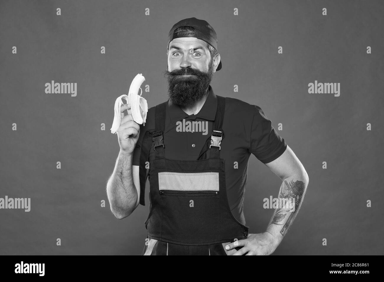 Mittagszeit. Bärtiger Mann in Uniform und Kappe, der Banane isst. Arbeiter Erbauer Mechaniker essen Banane. Frisches Obst. Gesundes Leben. Leckeres Essen. Bissen Sie Banane. Hungriger Flusspferd. Bevorzugter Snack. Lebensmittelgericht. Stockfoto