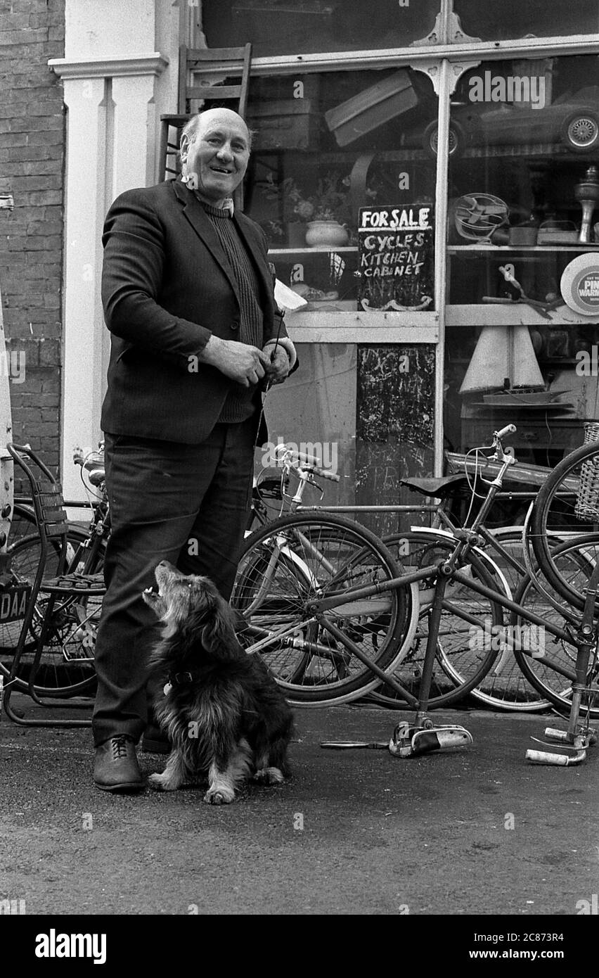 AJAXNETPHOTO. JANUAR 1975. SOUTHSEA, PORTSMOUTH, ENGLAND. - FAHRRADREPARATUREN - DER BESITZER DES SKIPPERS FAHRRADLADENS VOR DEM GELÄNDE MIT SEINEM HUND AN DER ECKE FONTWELL ROAD. FOTO: JONATHAN EASTLAND/AJAX REF:7504 202206 30 Stockfoto