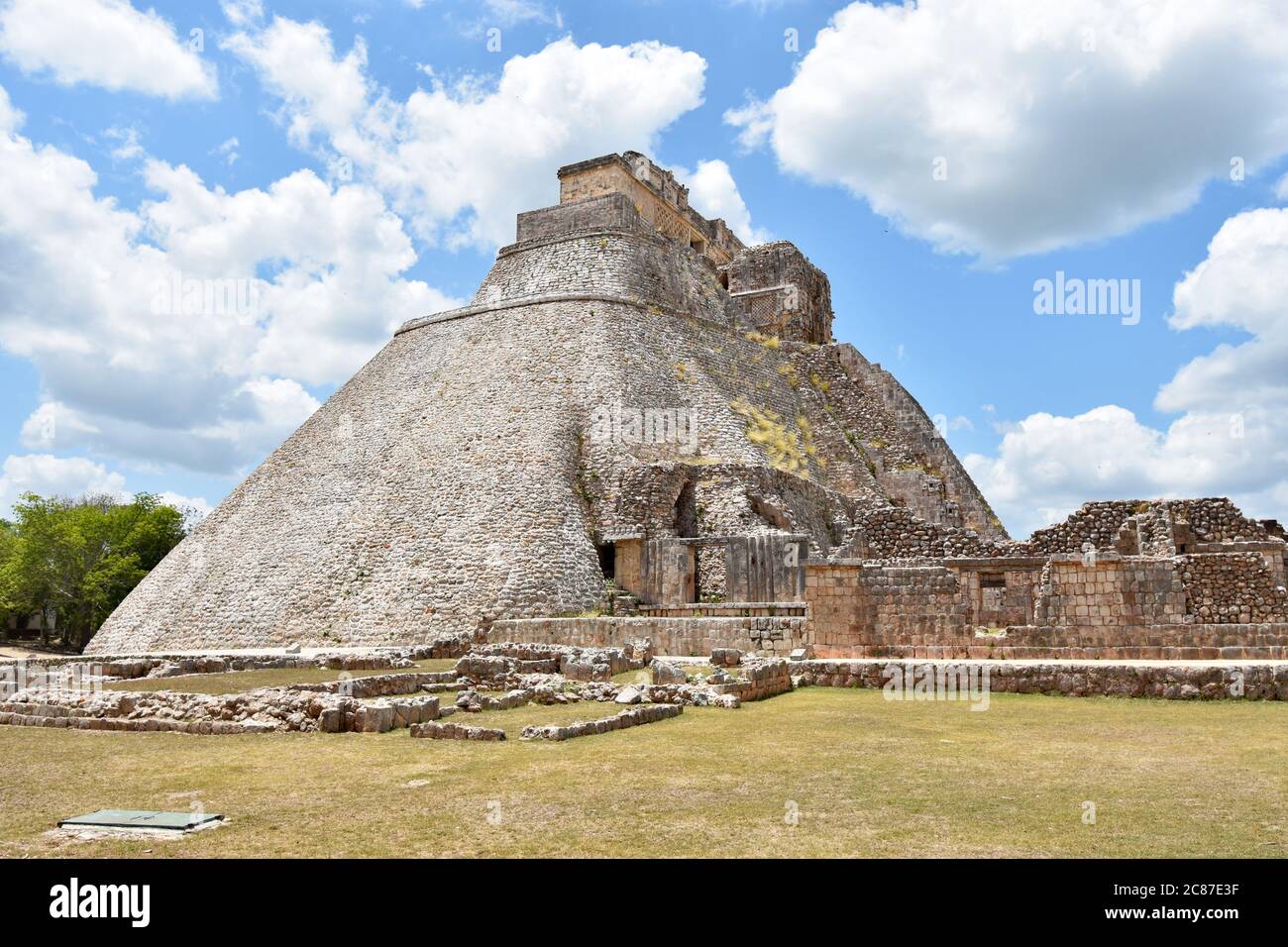 Die Pyramide des Magiers und die umliegenden Ruinen in der antiken Maya-Stadt Uxmal auf der Halbinsel Yucatan, Mexiko. Gelbfärbende Gräser und blauer Himmel. Stockfoto