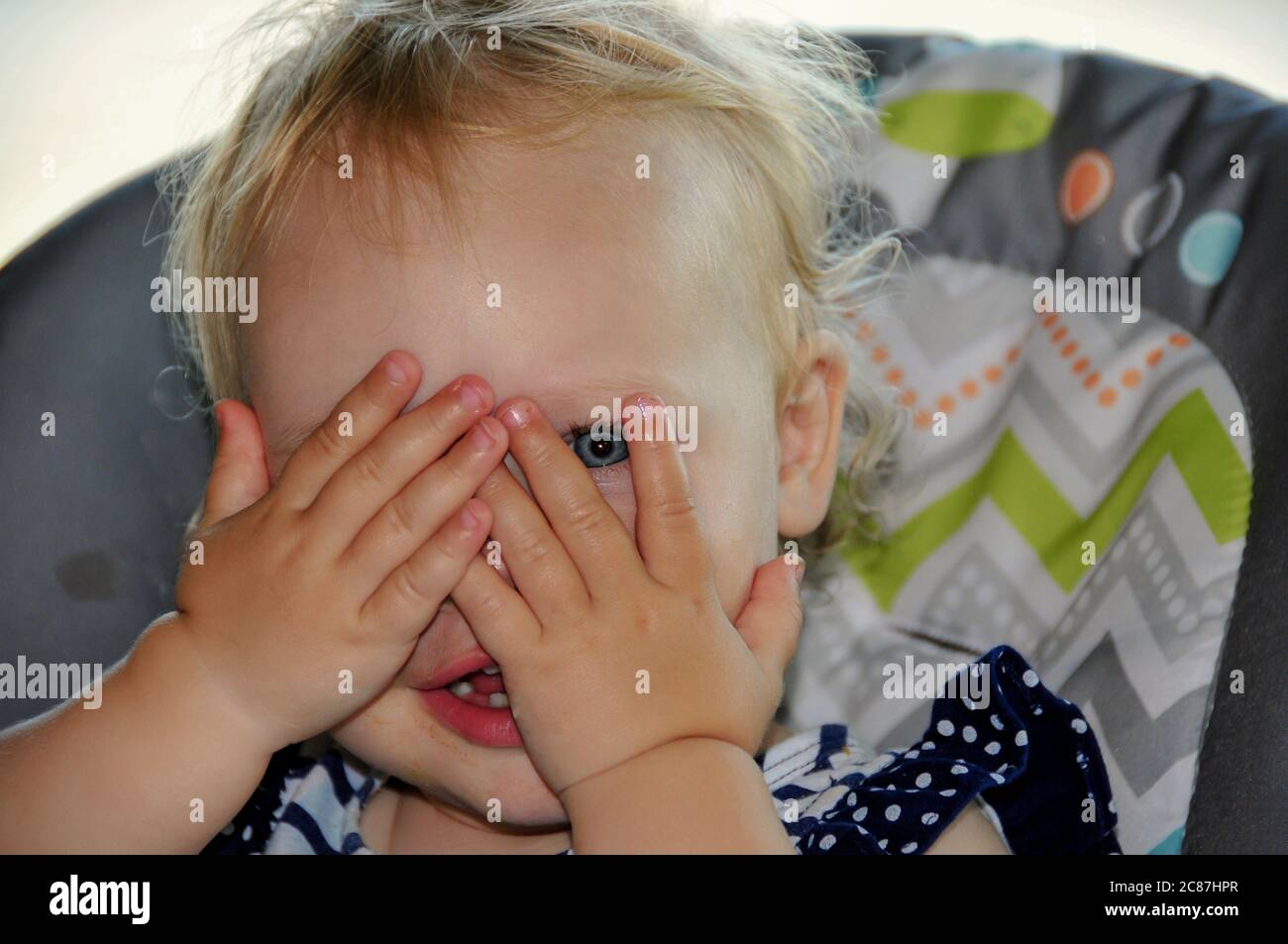 Liebenswert blonde kaukasische Baby spielen Peek-a-boo mit einem blauen Auge zeigt. Hände bedecken ihr Gesicht und sie guckt durch ihre Finger. Modell rel Stockfoto