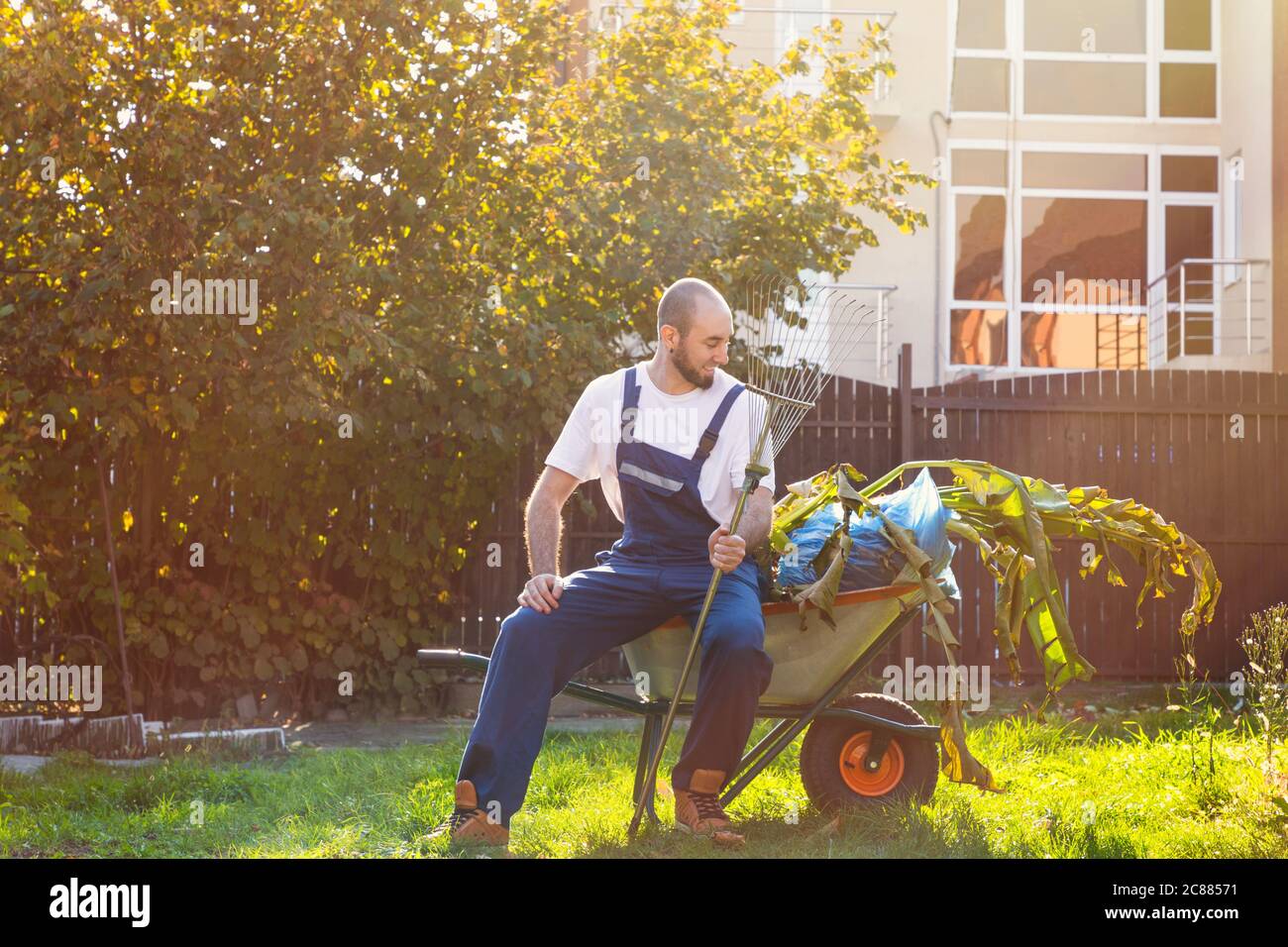 Der Gärtner sitzt auf dem Wagen mit Blättern. Die Sonne scheint hell. Lächeln und gute Laune. Gartenarbeit und Reinigung. Stockfoto