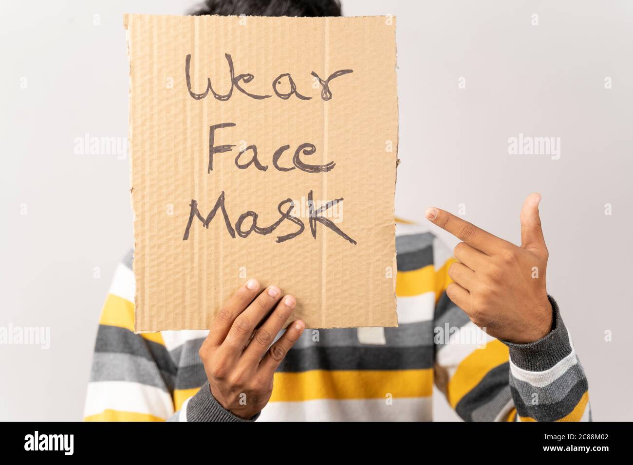 Junger Mann hält ein Pappschild, indem er sein Gesicht bedeckt, sagt, tragen Gesichtsmaske Nachricht auf isoliertem Hintergrund vor Coronavirus oder covid-19 zu schützen. Stockfoto