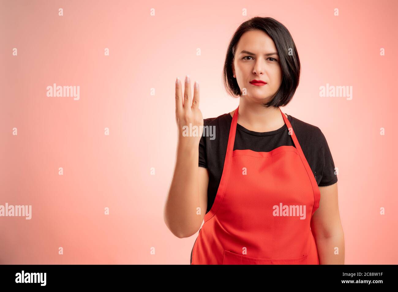 Frau beschäftigt im Supermarkt mit roter Schürze und schwarzem T-Shirt, zählen drei mit ihren Fingern isoliert auf rotem Hintergrund Stockfoto