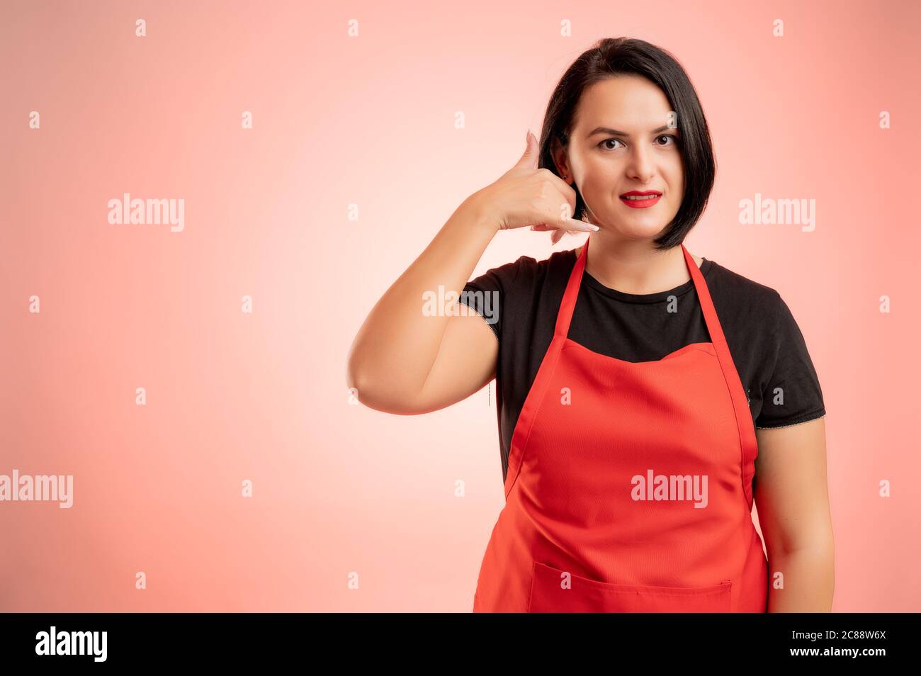 Frau im Supermarkt mit roter Schürze und schwarzem T-Shirt beschäftigt, zeigt Call me Geste isoliert auf rotem Hintergrund Stockfoto