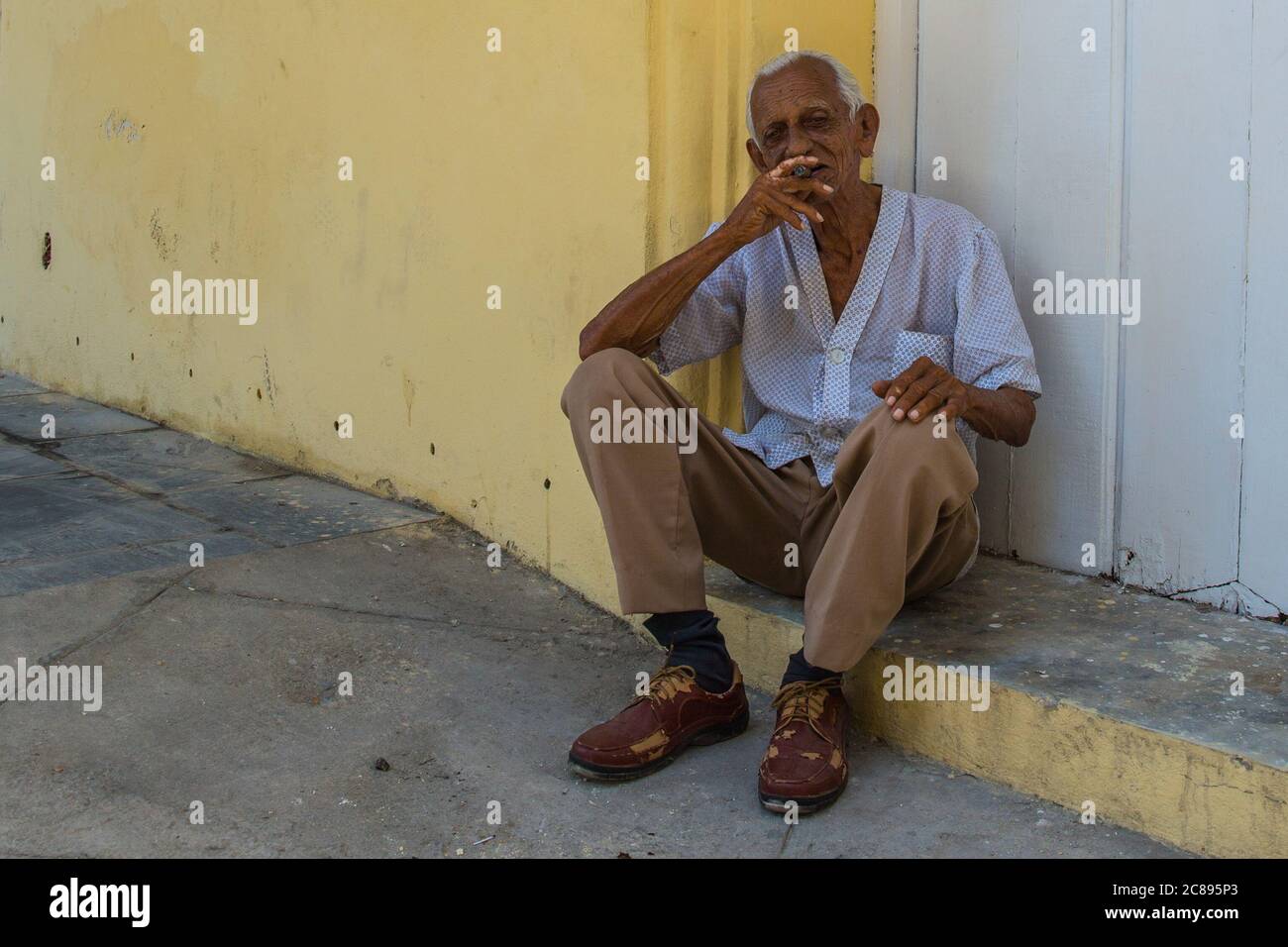 Havanna / Kuba - 04.15.2015: Ein alter Kubaner sitzt auf der Straße vor einem gelben Gebäude und raucht eine Zigarre in der Altstadt, Havanna, Kuba Stockfoto