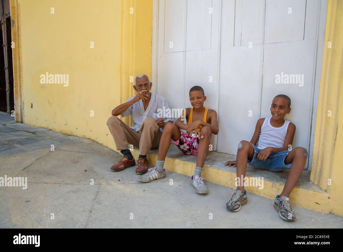 Havanna / Kuba - 04.15.2015: Alter Kubaner und Kinder sitzen auf der Straße vor einem gelben Gebäude; alter Mann raucht eine Zigarre in der Altstadt, Havanan Stockfoto