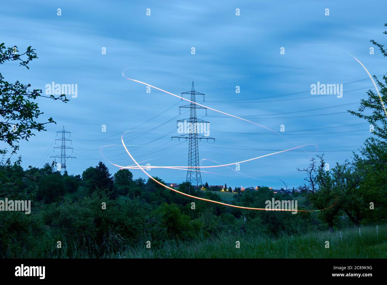 Großer Stromzonenmast auch Freileitungsmast steht hinter grüner Baumwiese, orangene Lichteffekte sind zu sehen. Deutschland. Stockfoto