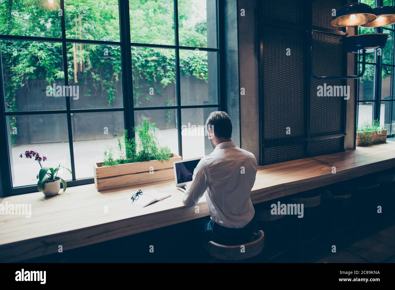 Rückansicht eines Journalisten ein stilvoller Typ, der eine Geschichte in einem Arbeitsraum in Coworking im Loft-Stil schreibt, gut gekleidet, neben dem Fenster sitzend mit Blick auf den Garten Stockfoto