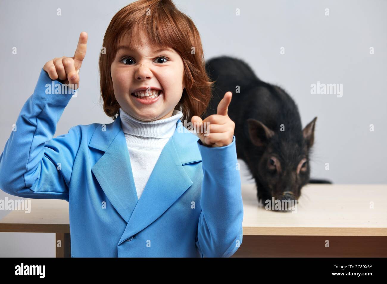 Indoor-Porträt von kleinen rothaarigen Jungen in blauer Jacke mit seinem exotischen Haustier - kleine schwarze Schwein, hübsches Kind Blick auf die Kamera und lächelnd. Stockfoto