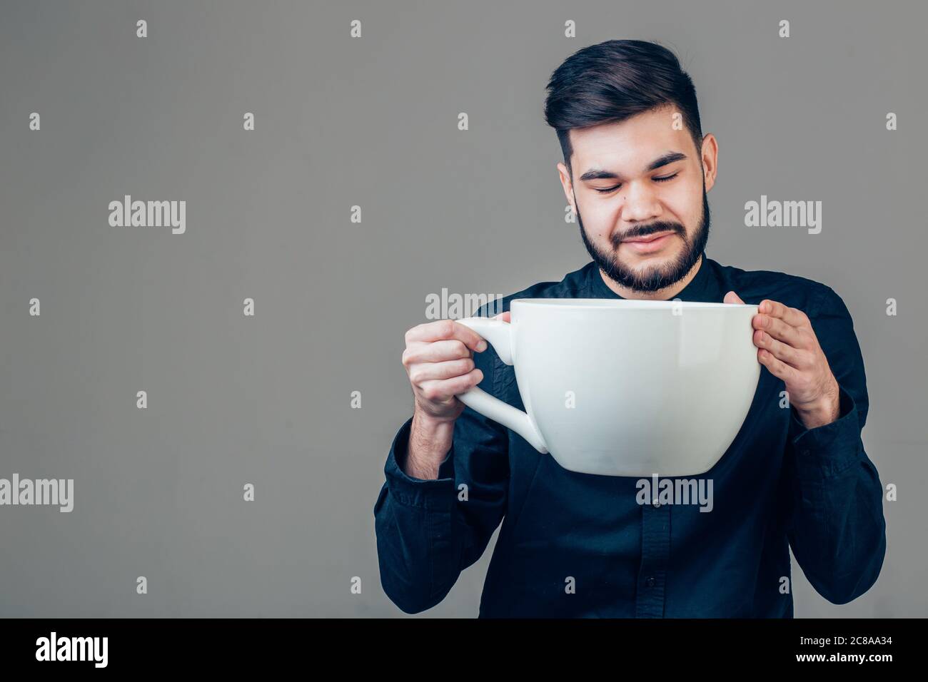 Junge glückliche Geschäftsmann hält eine lustige riesige und überdimensionale Tasse schwarzen Kaffee in Koffein sucht Konzept isoliert auf gleichem Hintergrund Stockfoto