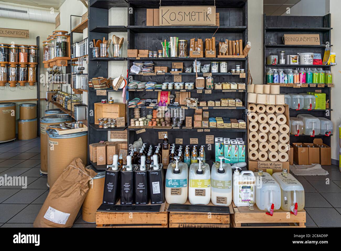 Ein Schild mit der Aufschrift „Kosmetik“ hängt über Regalen mit Zahnbürsten, Seifen und Verpackungsmaterial im Zero-Waste-Laden Basel Unverpackt in der Feldbergstrasse in Basel, Schweiz. Vorne stehen Kanister sowie nachfüllbare Seifen- und Duschgelspender, daneben gestapelte Rollen Bambus-Toilettenpapier neben einem Regal mit Reinigungsprodukten rechts. Stockfoto