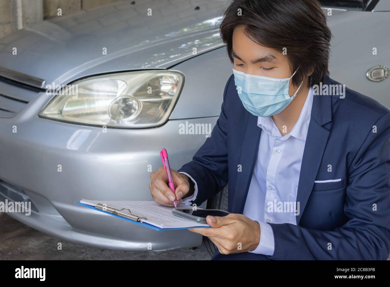 Sitzender asiatischer Versicherungsagent oder Versicherungsagentur in Anzug Wear Mask Überprüfung Auto Crash Daten Bericht von Unfall für Anspruch in Garage Stockfoto