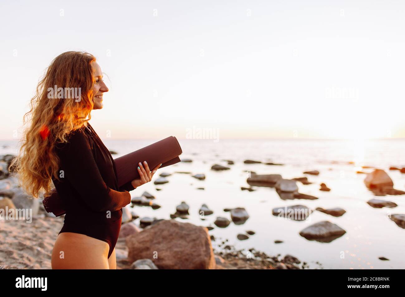 Junge schöne Frau mit Yoga-Matte stehen am Strand im Sommer bei Sonnenuntergang und bereit für das Training. Stockfoto