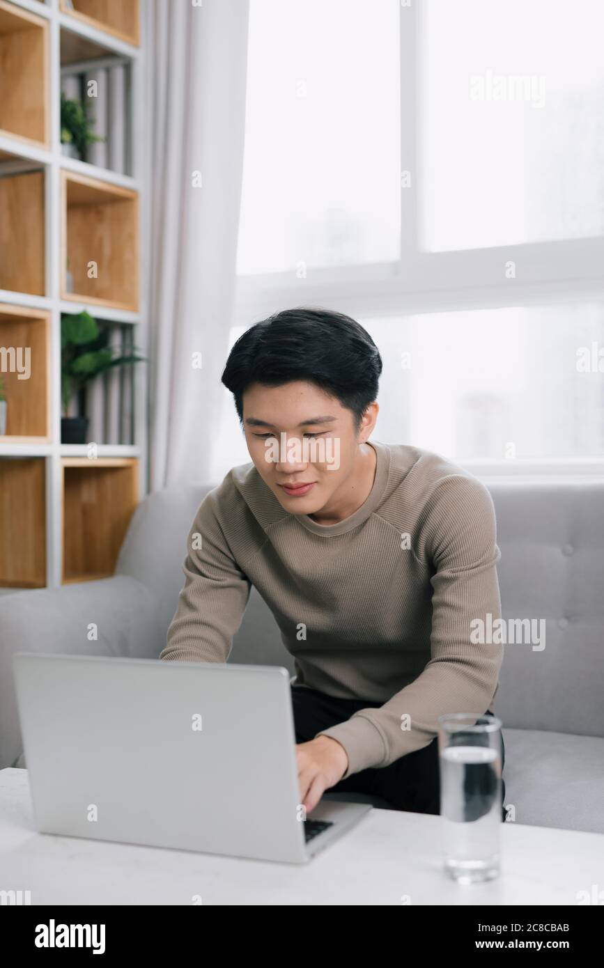 Junge attraktive lächelnden Kerl ist Browsen mit seinem Laptop, zu Hause sitzen auf der gemütlichen beige Sofa zu Hause, Legere Outfit Stockfoto