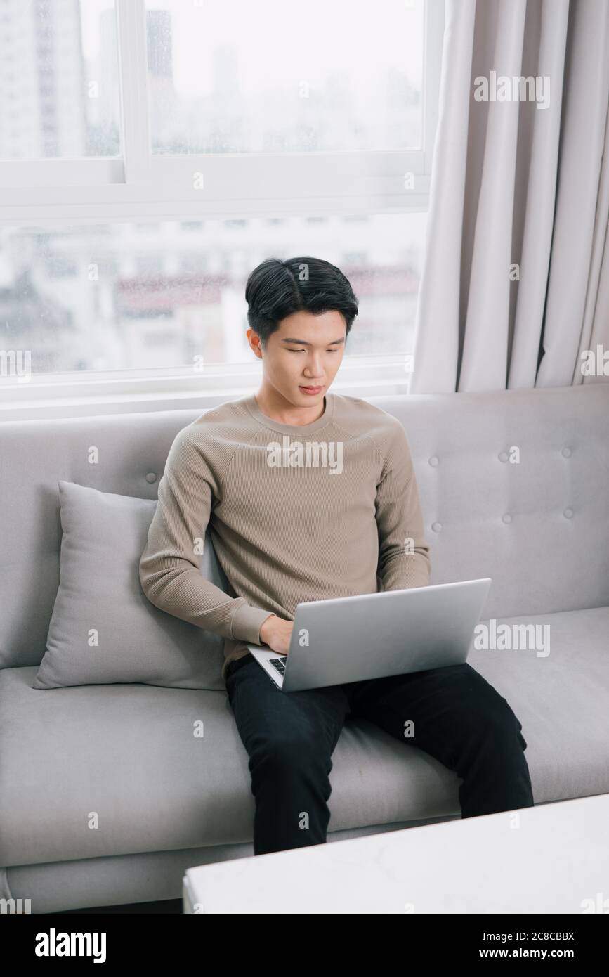 Junge attraktive lächelnden Kerl ist Browsen mit seinem Laptop, zu Hause sitzen auf der gemütlichen beige Sofa zu Hause, Legere Outfit Stockfoto