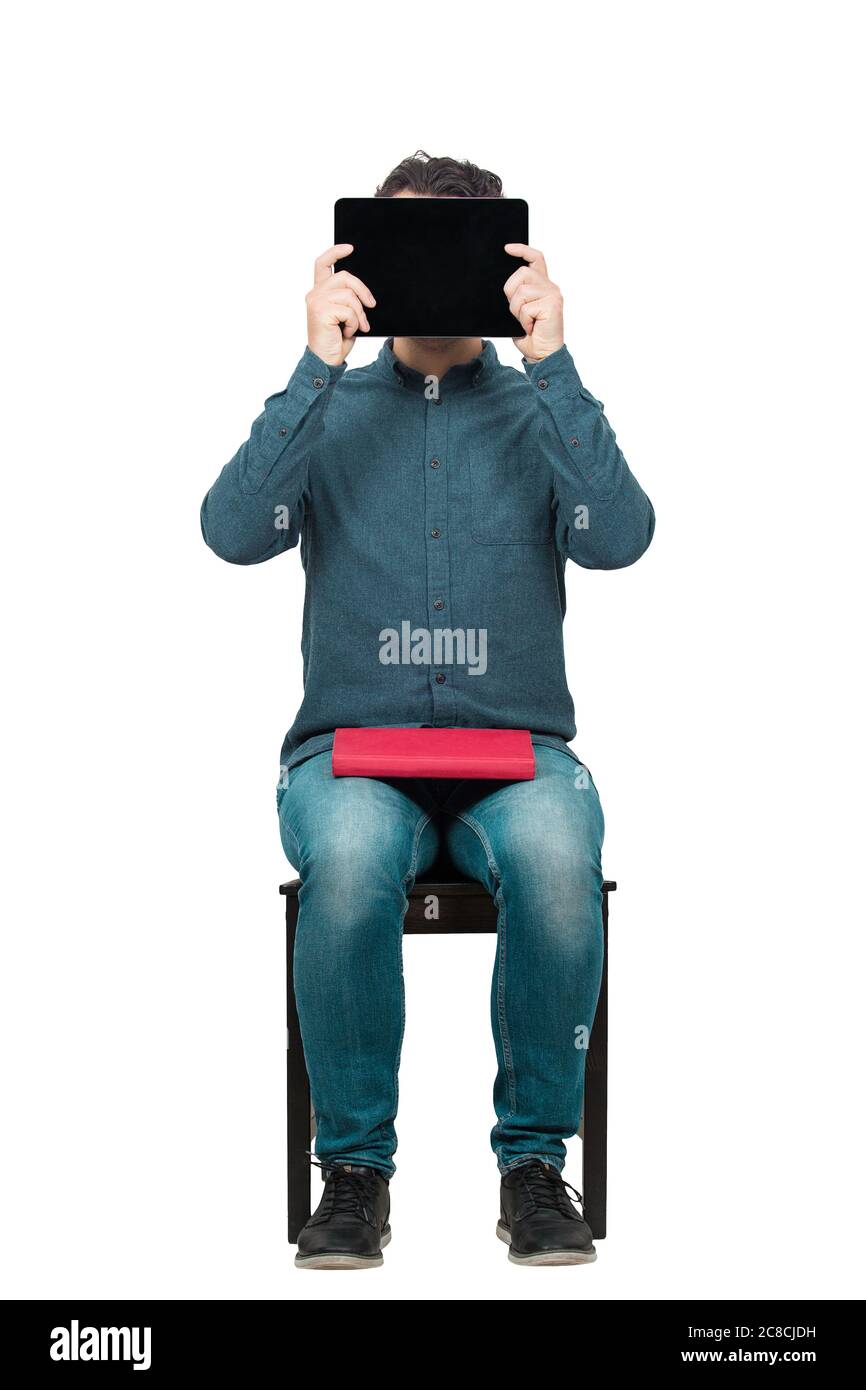 Ein junger Student, der auf einem Stuhl sitzt, bedeckt seinen Kopf mit einem PC-Tablet, während er ein rotes Buch auf den Knien hält. Modernes Bildungskonzept. Studieren und Arbeiten Stockfoto