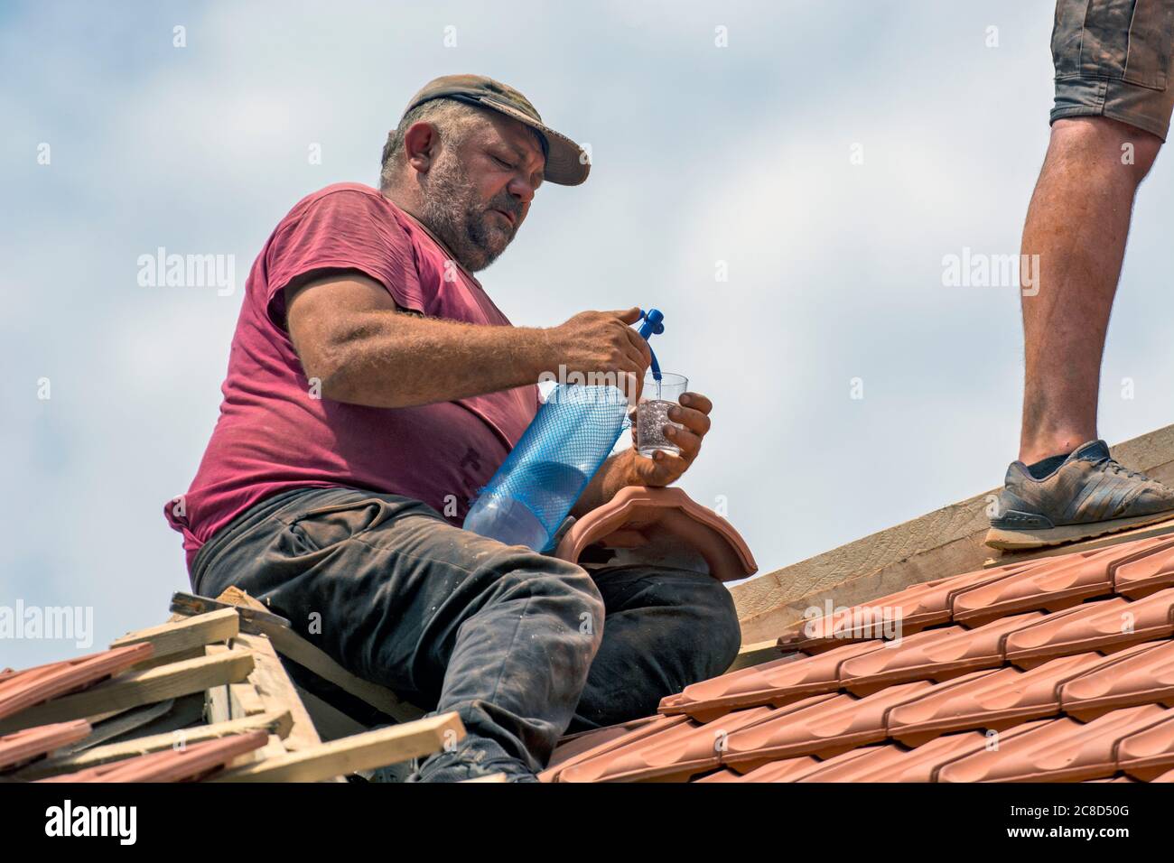 Zrenjanin, Serbien, 23. Juli 2020. Meister arbeiten auf dem Dach eines Privathauses, um eine alte Fliese zu ersetzen. Einer der Meister erfrischt sich mit Stockfoto