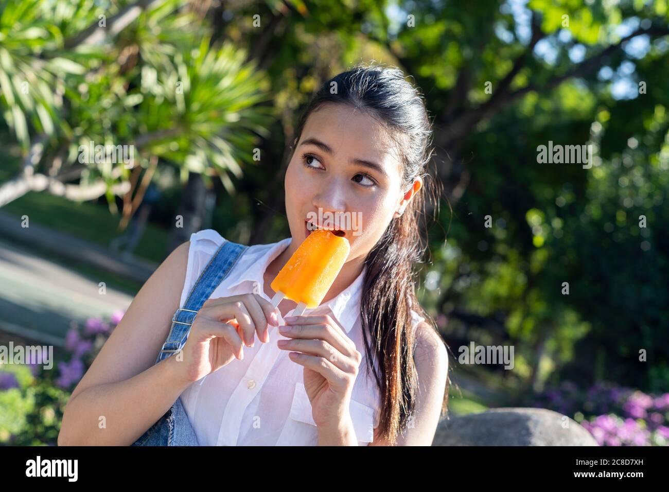 Pferdeschwanz Mädchen in Denim Overalls und weißen Hemd beißen ein oranges Eis im Park an sonnigen Tag. Stockfoto