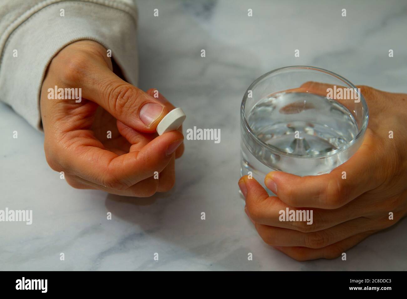 Eine kaukasische Frau hält eine weiße Pille in der einen Hand und hält ein Glas Wasser fest. Sie ist dabei, die Pille einzunehmen. Stockfoto