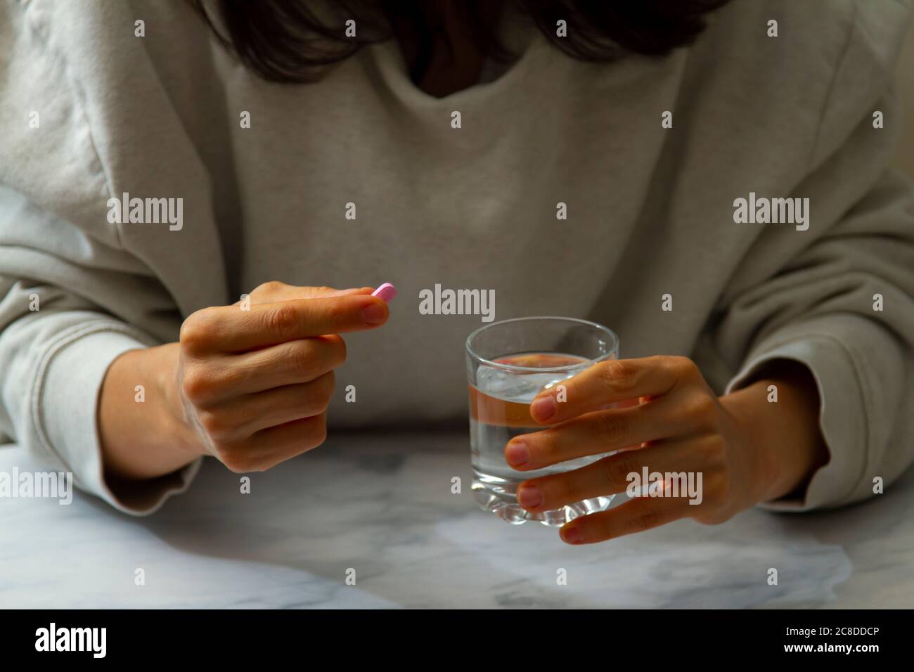 Eine Frau wird gesehen, wie sie eine Pille in der einen Hand und ein Glas Wasser auf der anderen Hand hält. Sie ist dabei, die Medikamente mit einem Schluck Wasser einzunehmen. Stockfoto
