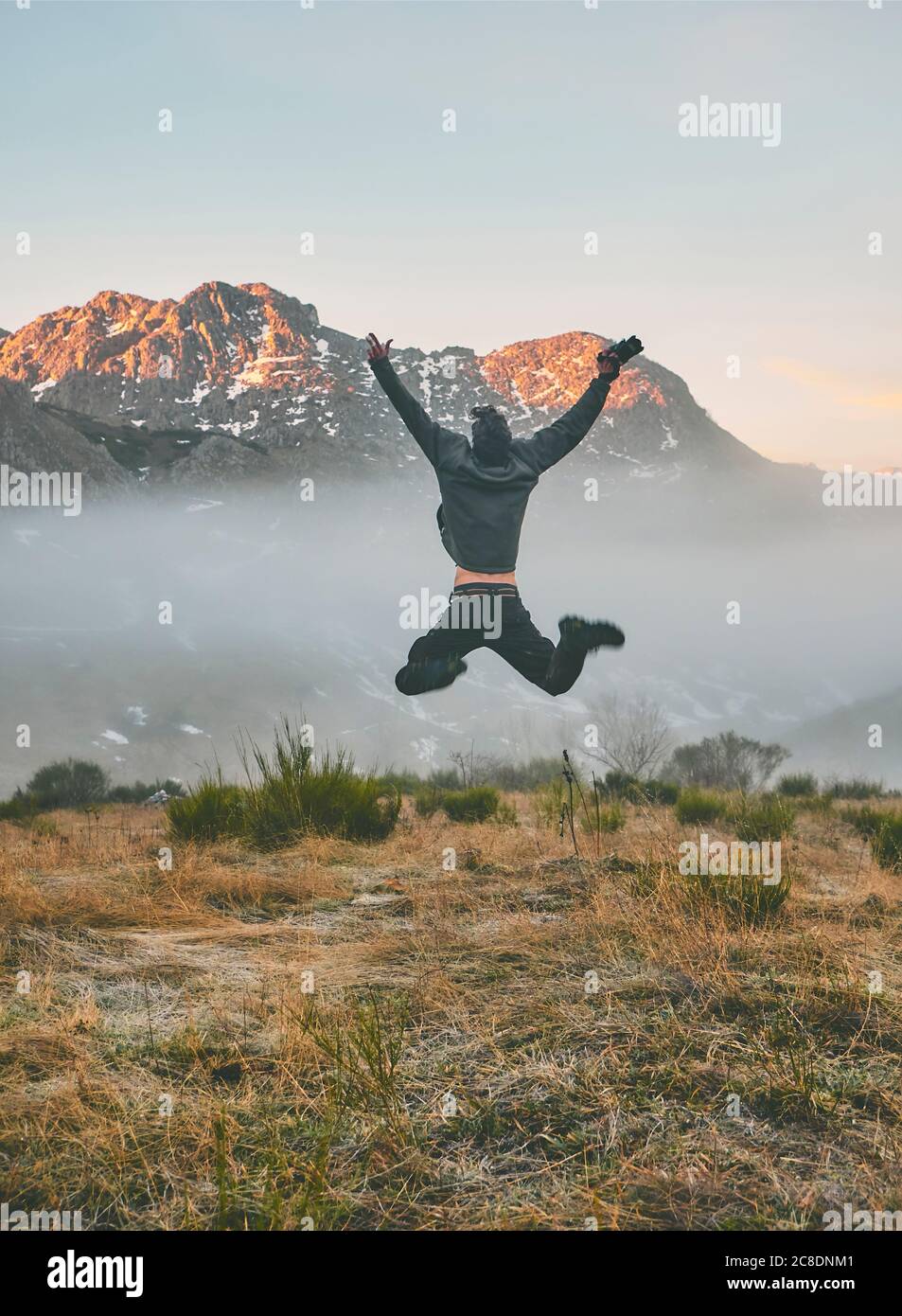 Junger männlicher Tourist mit ausgestreckten Armen, der während des Sonnenaufgangs auf den Berg springt, Leon, Spanien Stockfoto